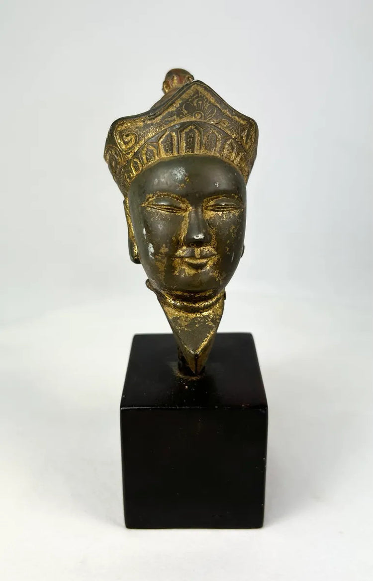 Fragmentary Tibetan Buddha head in gilded bronze, 18th–19th century, featuring serene expression, half-closed eyes, elongated ears, and ornate crowned headdress, displayed on a wooden base.