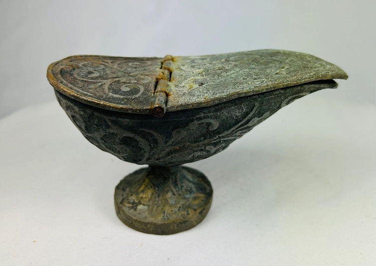 Three-quarter angle of ancient bronze oil lamp showing hinged lid, swirling engraved motifs, and oxidation from centuries of burial.