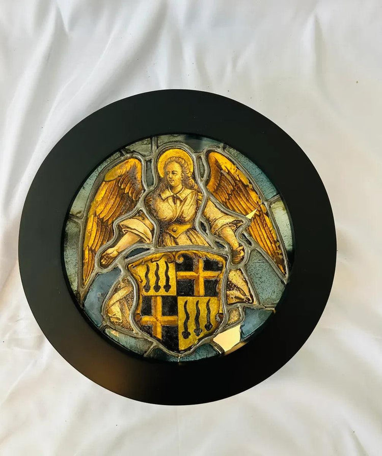 16th-century Swiss stained glass panel featuring a nimbate angel with outstretched wings holding a heraldic shield adorned with golden crosses and ermine motifs, displayed in a circular black frame against a white background.