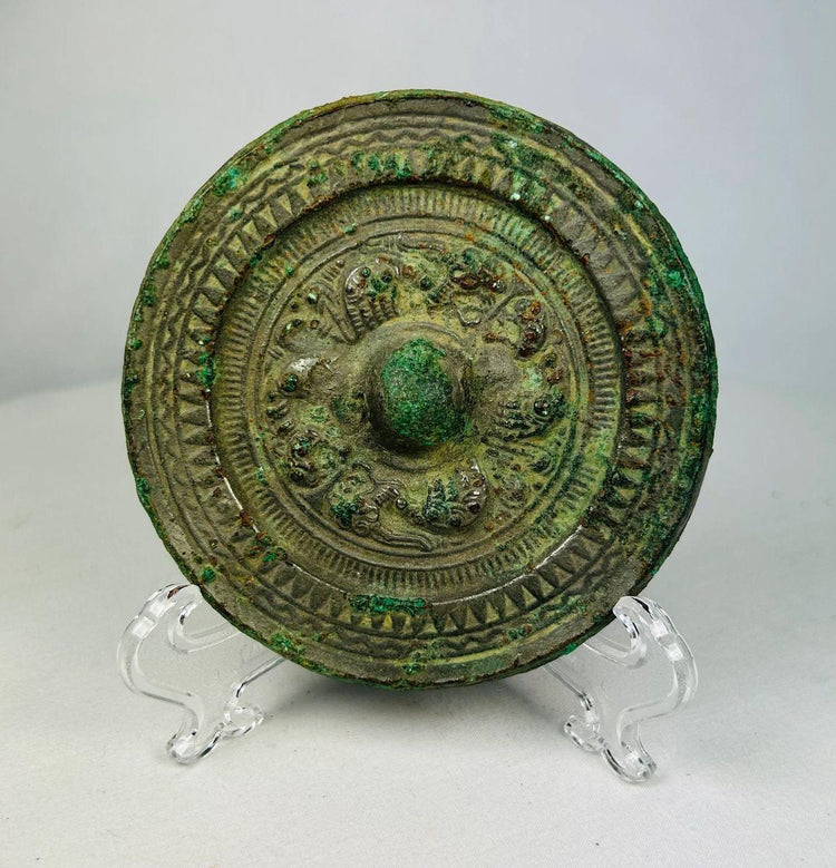Ancient Han Dynasty bronze mirror with green patina, circular motifs, and raised central knob displayed on a clear stand.