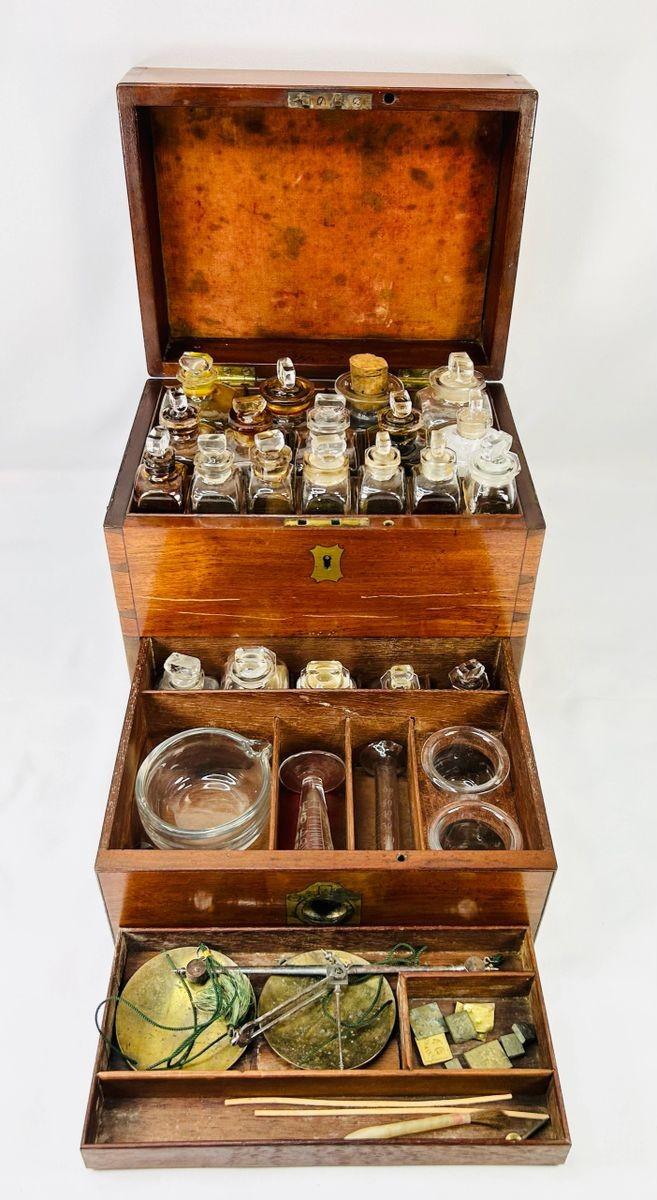 Antique Victorian apothecary box with drawers open, displaying glass bottles, brass scales, mortar and pestle, and chemist’s tools — 19th-century England.