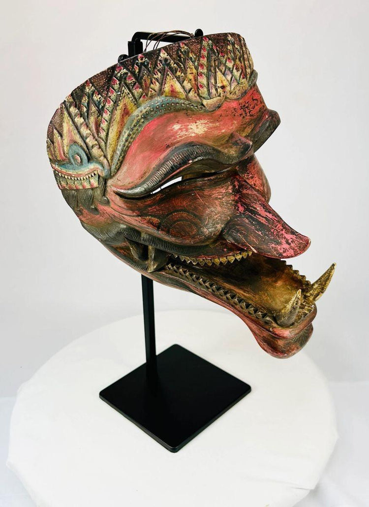 Hand-carved Javanese Cakil mask with aged painted details, displayed at an angle on a black stand.