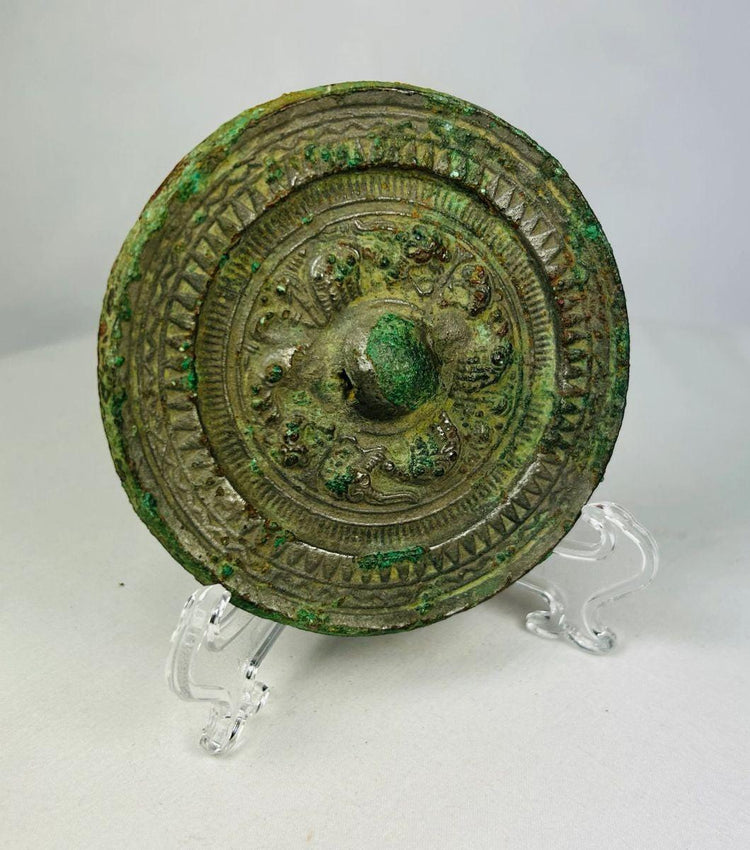Han Dynasty bronze mirror with intricate concentric designs and green patina displayed on a clear stand.