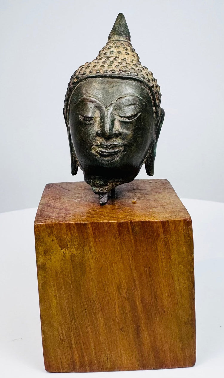 Thai bronze Buddha head displaying arched brows, softly curved lips, and elongated earlobes above a warm brown wooden block.