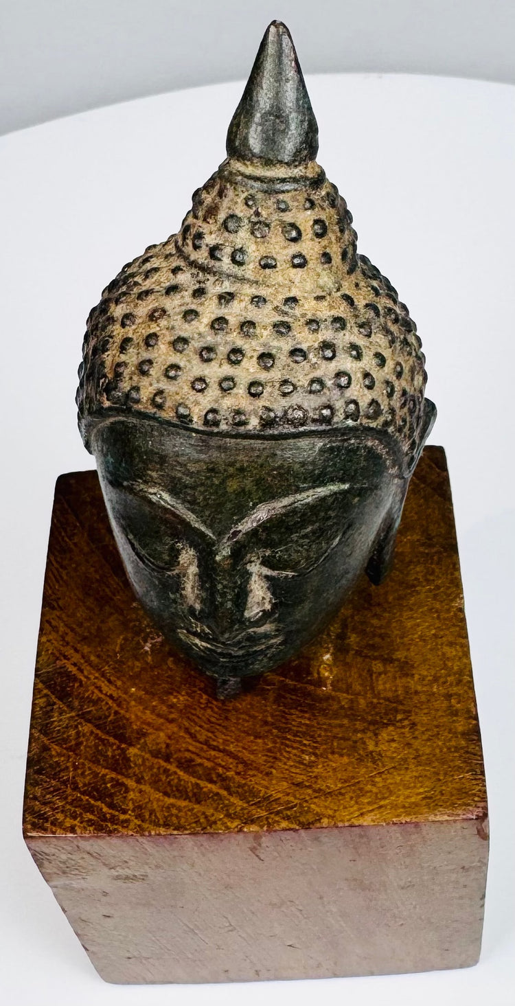 Bronze Buddha head highlighting the pointed topknot, lighter textured hair, and arched brows above half-closed eyes.