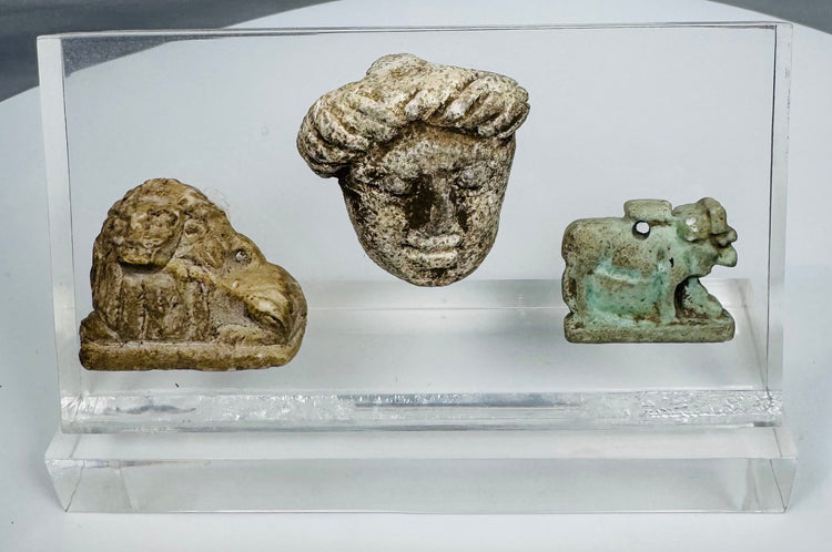 Archaeological artifact group including bovine-form Hathor amulet, miniature lion sculpture, and Hellenistic-style head.