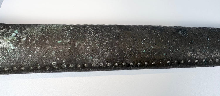 Metal sword sheath with red and dark cabochon inserts and green surface corrosion.