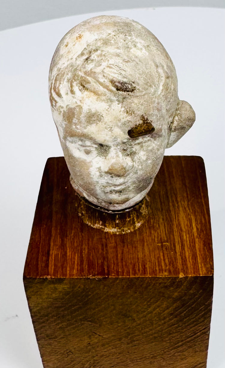 Ancient terracotta head showing sculpted hair, recessed eyes, and a small mouth above its warm brown wooden display mount.
