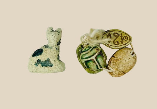 Ancient Egyptian faience cat amulet with traces of blue-green glaze, shown alongside three scarabs in varied materials, on a white background.