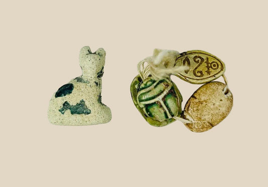 Ancient Egyptian faience cat amulet with traces of blue-green glaze, shown alongside three scarabs in varied materials, on a white background.