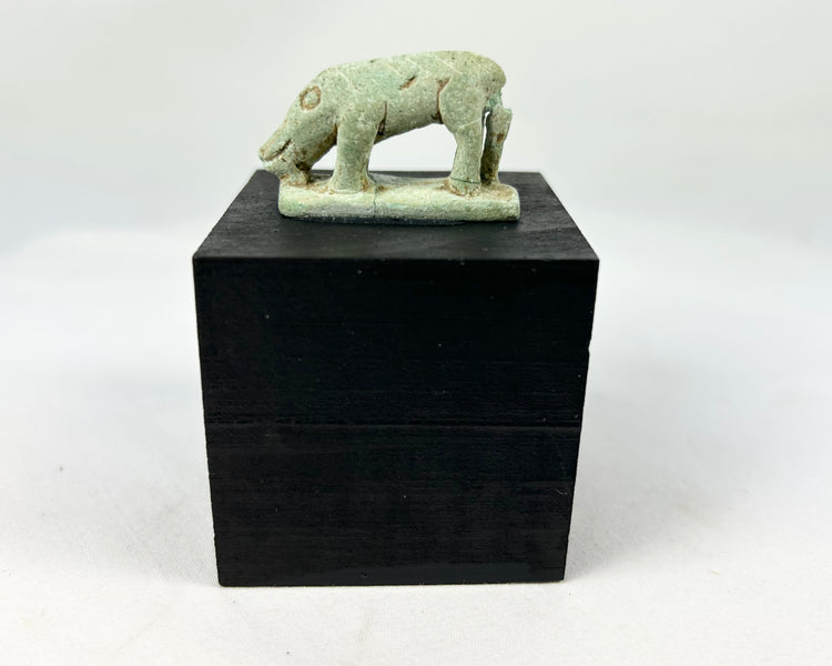 Miniature Egyptian faience shrew amulet with elongated snout, ring-and-dot eye, four legs, and rear support strut on rectangular plinth, traces of green glaze and mineral accretions, Late Period–Ptolemaic Egypt, ca. 664–30 BCE, displayed on modern wood block.