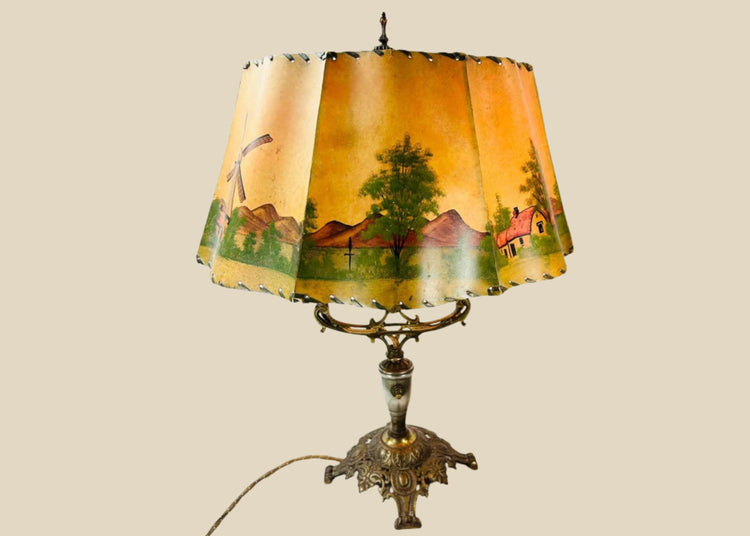 Antique Art Nouveau table lamp, circa 1920s, with ornate cast metal base and original hand-painted parchment shade depicting cottages, windmills, and trees, a decorative European lighting piece with warm pastoral charm.
