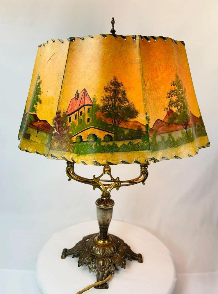 Antique Art Nouveau table lamp, circa 1920s, with ornate cast metal base and original hand-painted parchment shade depicting cottages, windmills, and trees, a decorative European lighting piece with warm pastoral charm.