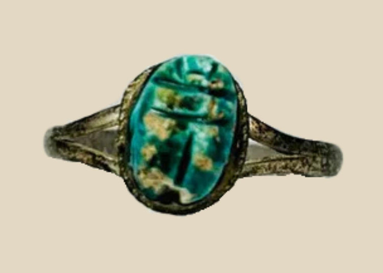Ancient Egyptian scarab ring with turquoise-green faience scarab set in bronze band, displayed on a clear stand against a white background.