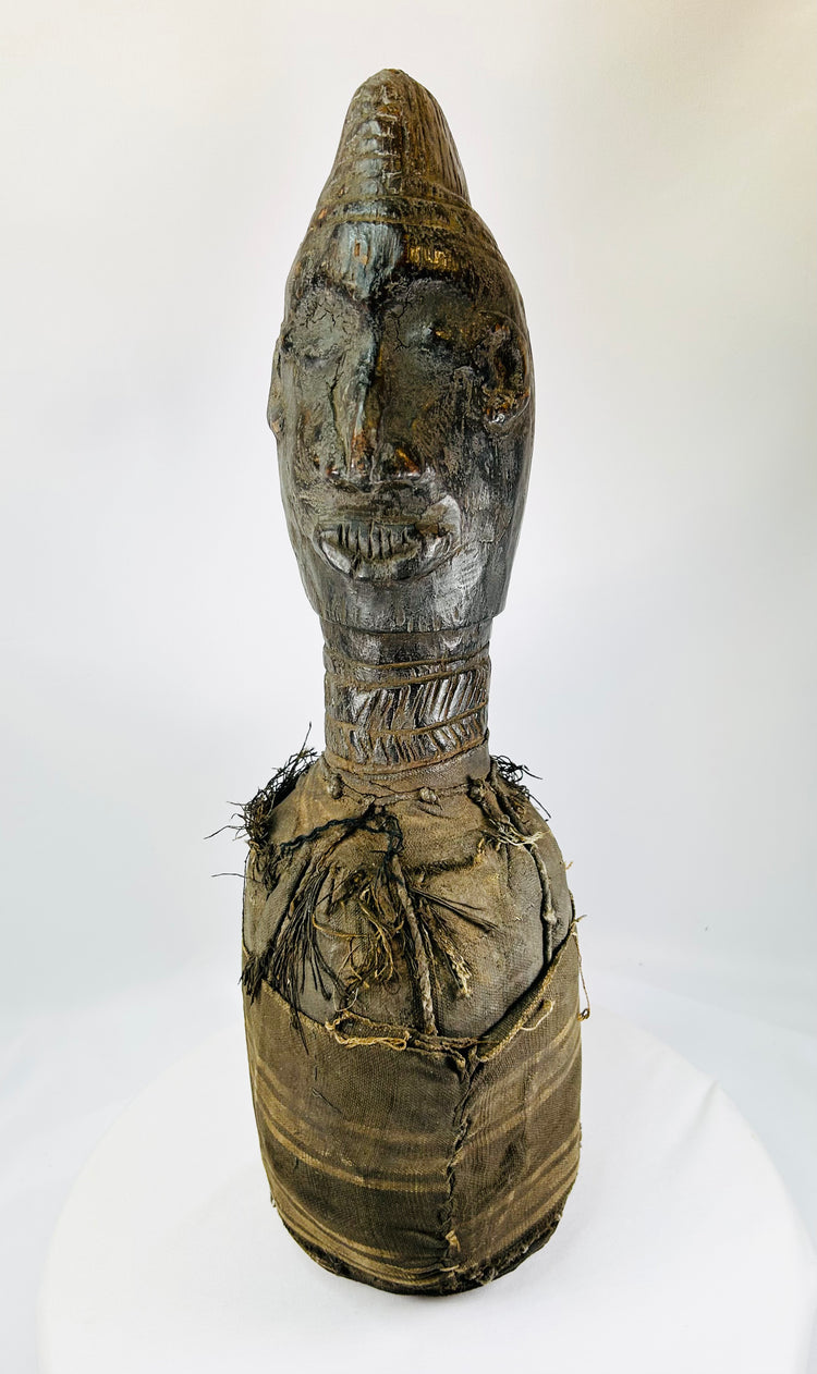 Hand-carved Central or West African wooden figure with elongated facial features, natural fiber torso wrapping, and traces of organic pigments, standing on a white background.