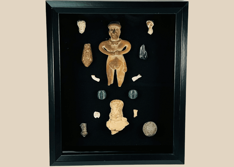 Framed Pre-Columbian artifact shadow box with terracotta, stone, and shell pieces behind glass, showing light reflections on the display surface.