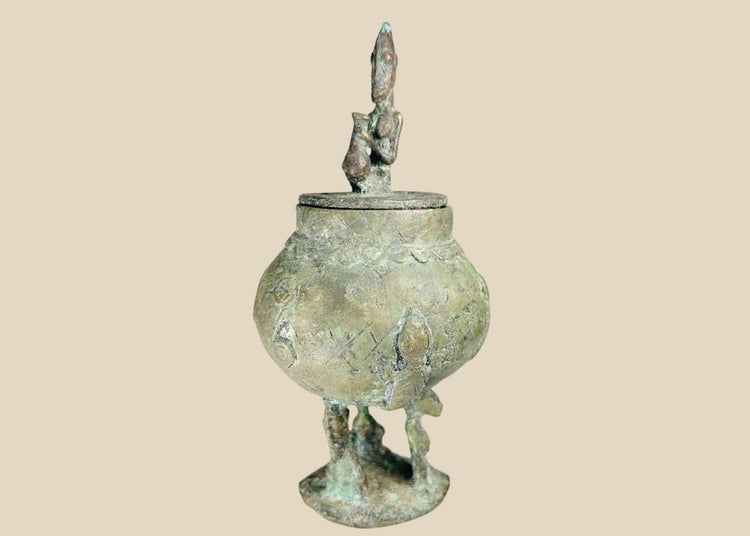 19th-century Dogon bronze container from Mali with bulbous body, engraved motifs, anthropomorphic legs, and a sculpted human figure on the lid, used in ritual and healing practices.