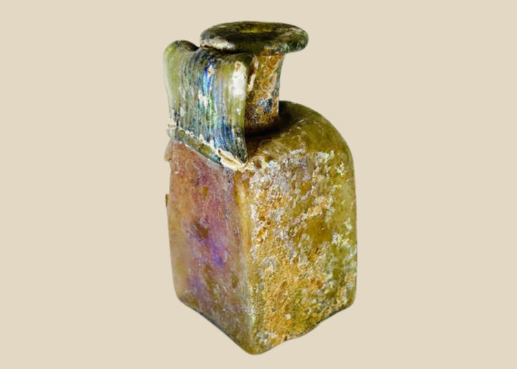 Early Imperial Roman mold-blown glass bottle from the 1st Century CE, likely Rhineland. Square translucent body with cylindrical neck, flaring folded rim, and wide ribbed “celery handle.” Base bears four-leaf maker’s mark. Developed iridescent patina reflects age and Roman glassmaking innovation, used for oils, perfumes, or wine.