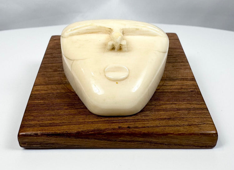 Abstract tribal-inspired bone face carving with closed eyes and simplified features, presented on a polished hardwood plaque.
