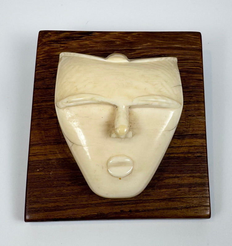Decorative bone spirit head carving with geometric contours and smooth polished finish, mounted against a rich hardwood display plaque.