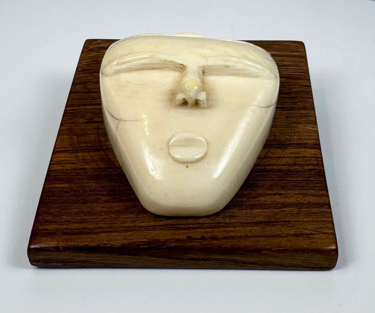 Vintage bone relief carving of a human spirit face with closed eyes and triangular form, mounted on a polished wooden plaque.