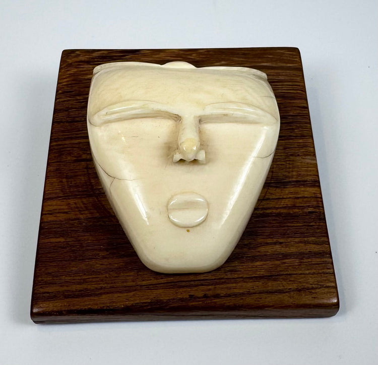 Mid-20th century bone carving depicting a stylized spirit face with an elongated nose, rounded lips, and smooth ivory-toned surface displayed on a wooden plaque.