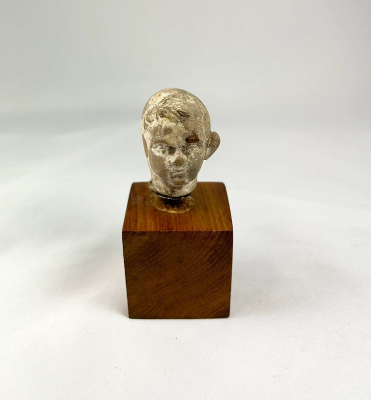 Small terracotta head fragment of a youthful male from the Hellenistic or early Roman period, with delicate features, subtle smile, and natural patina, mounted on a wooden display base.