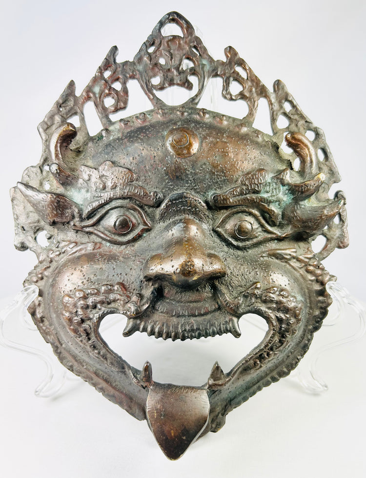 Late 19th–early 20th century Himalayan bronze or brass ritual mask, depicting fierce protective deity with wide eyes and grimacing mouth, traces of pigment, patina, used in Buddhist or Hindu ceremonies