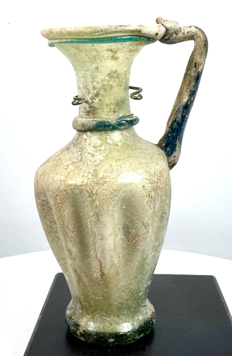 Late Roman glass jug with an elegant ovoid body, flared trumpet-shaped mouth, curved handle, and extensive ancient surface patination.