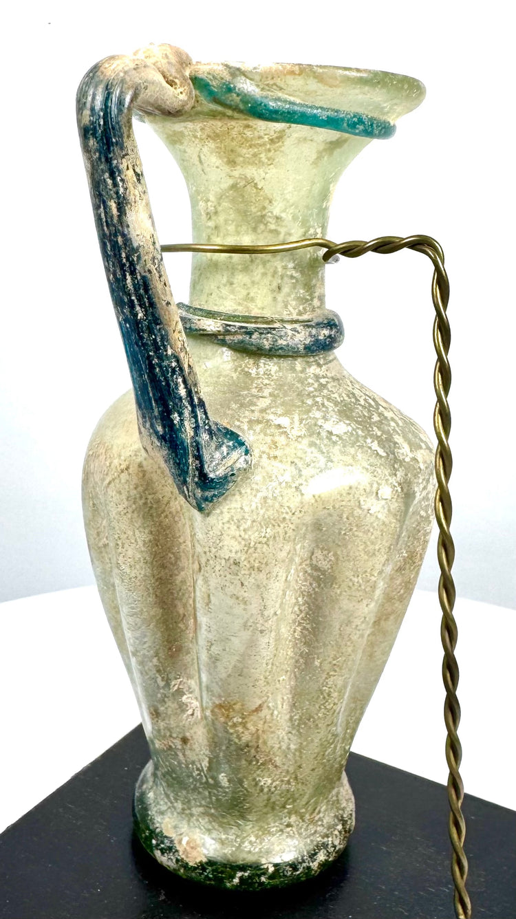 Ancient Roman glass pouring vessel with a gracefully curved handle, applied glass collar, and richly textured archaeological surface deposits.