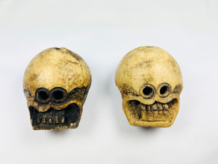 Front view of two Tibetan Citipati ritual beads carved from human femur bone, showing skull faces and suspension holes on a white background.