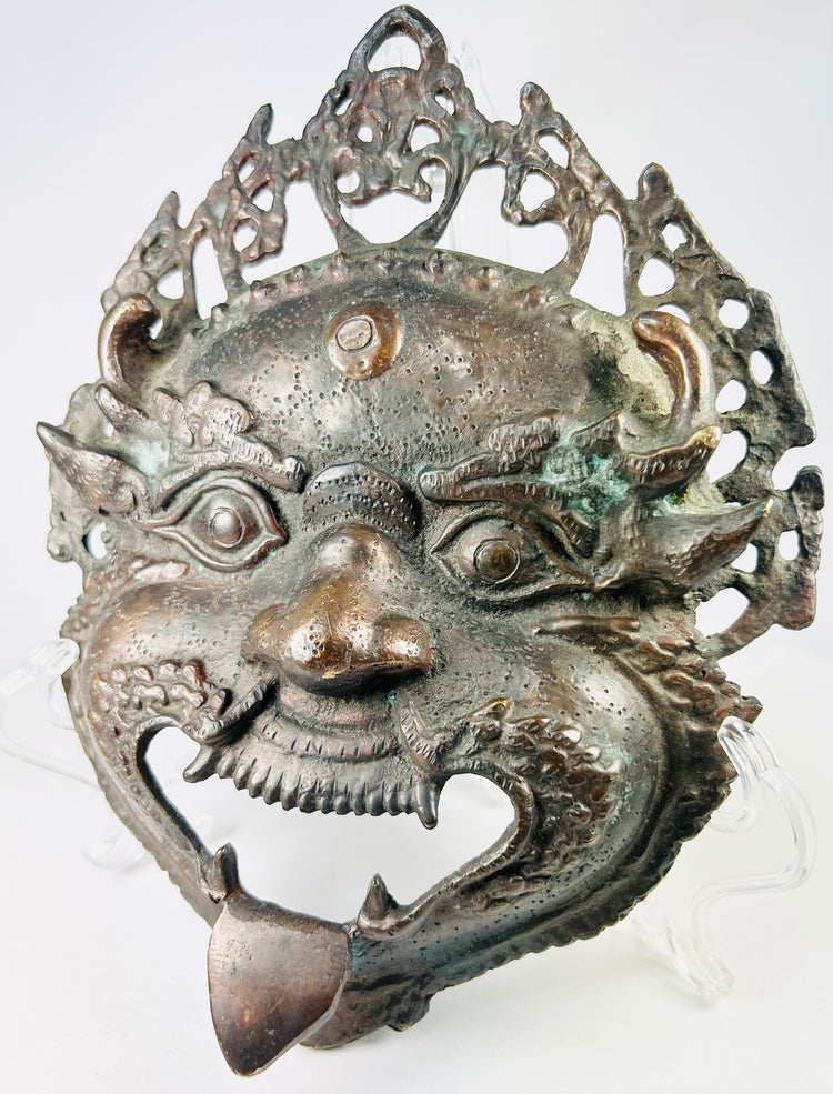 Late 19th–early 20th century Himalayan bronze or brass ritual mask, depicting fierce protective deity with wide eyes and grimacing mouth, traces of pigment, patina, used in Buddhist or Hindu ceremonies
