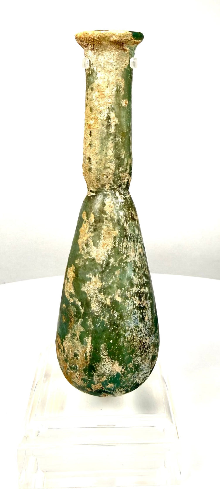 Ancient Roman unguentarium displaying earthy beige deposits and irregular weathering across its green glass surface.