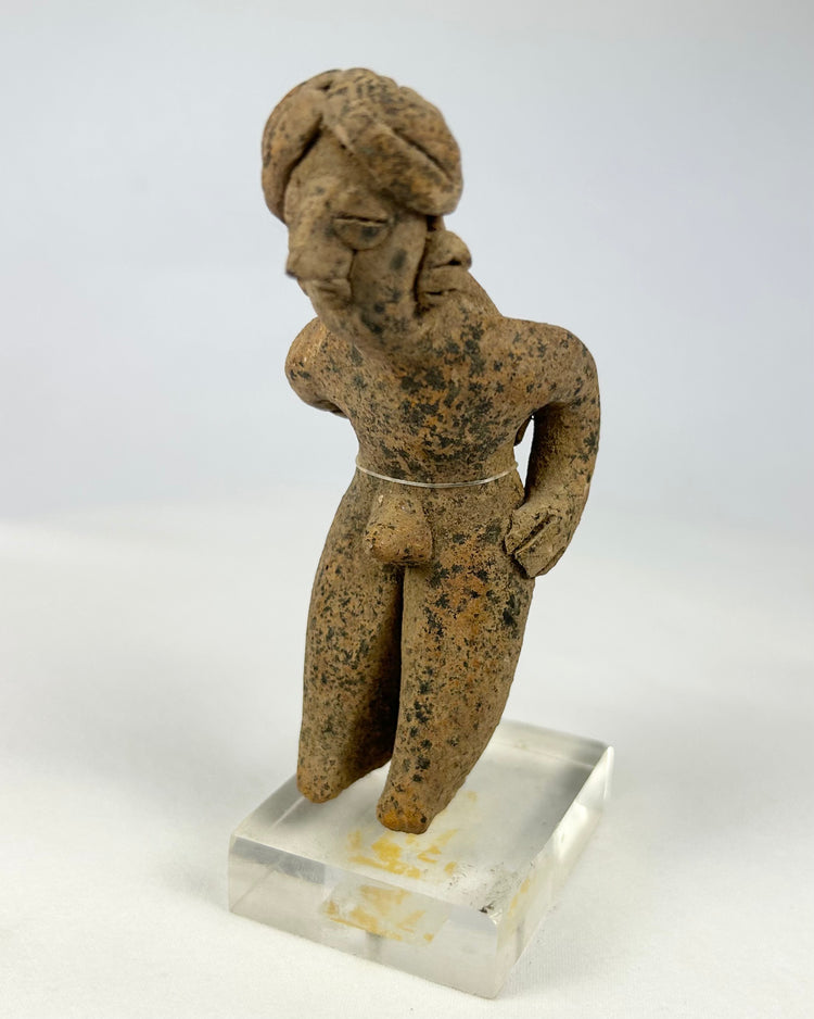 Pre-Columbian West Mexican terracotta standing figure, circa 300 BCE–500 CE, hand-modeled with tall turban-style headdress, stylized proportions, and surface encrustations, mounted on acrylic display base
