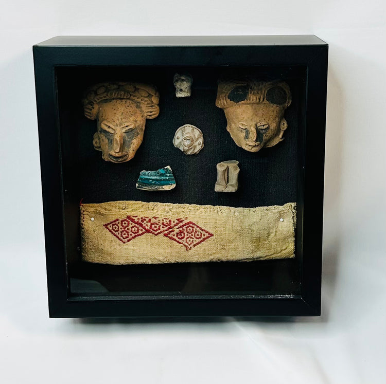 Framed display of ancient terracotta masks, carved ornaments, and Pre-Columbian woven textile