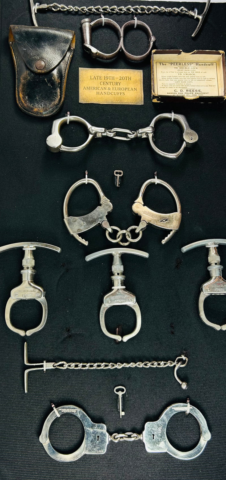 19th–20th century antique handcuffs, shackles, keys, and leather pouch arranged in a framed historical restraint collection on dark fabric background.