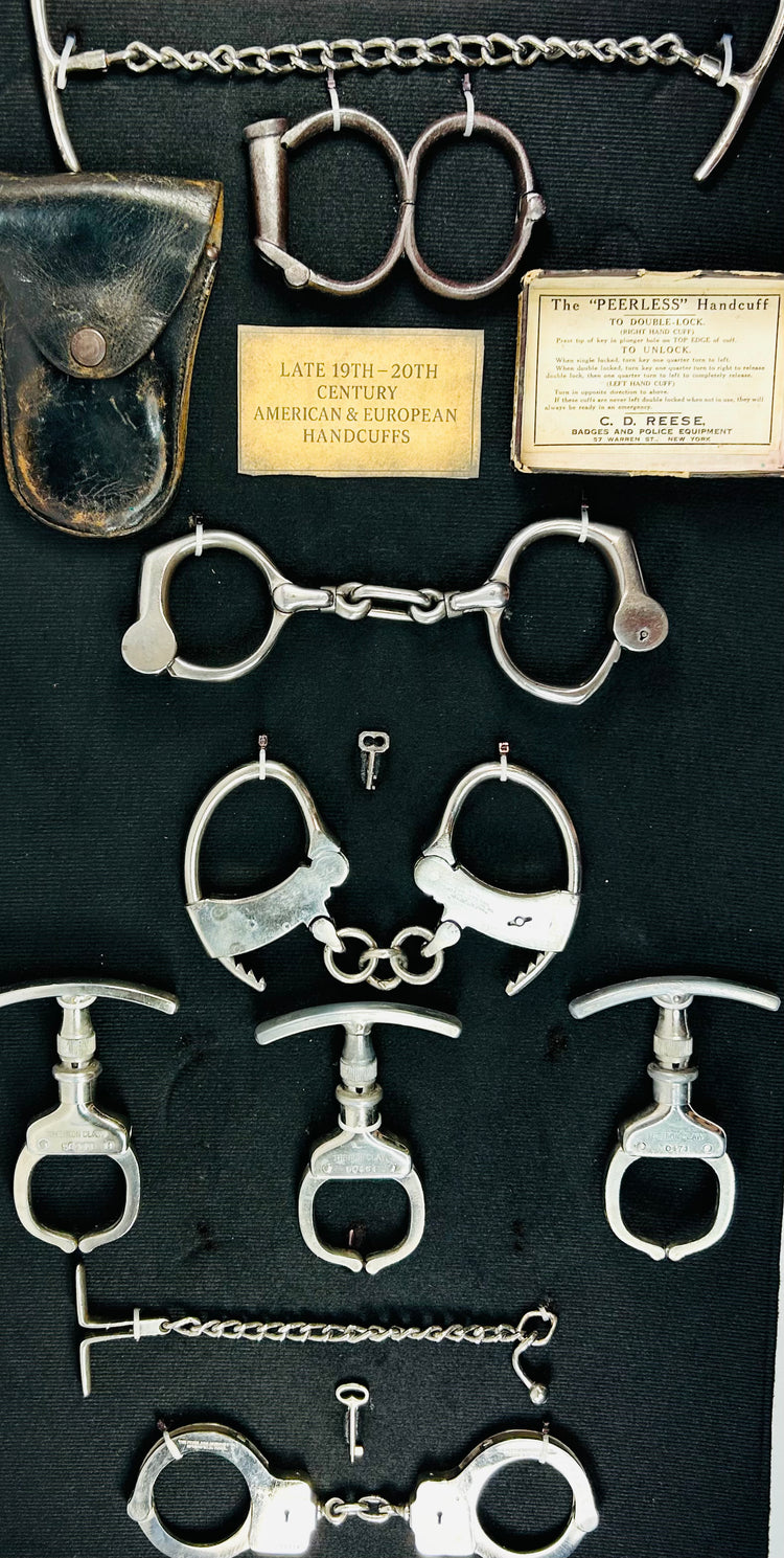 Antique handcuffs, shackles, keys, and leather pouch displayed with identification plaques in a framed 19th–20th century restraint collection on dark fabric.