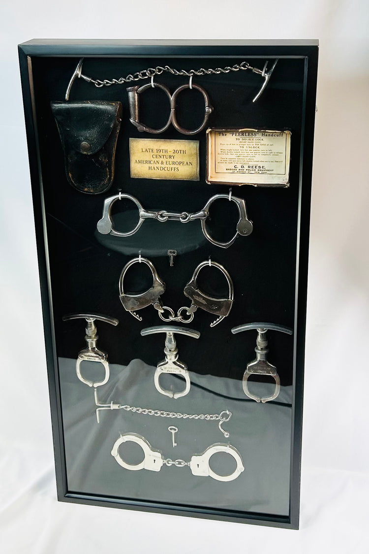 Framed display of antique handcuffs, shackles, and restraint devices from the 19th–20th century, mounted in a glass shadow box with plaques and keys.