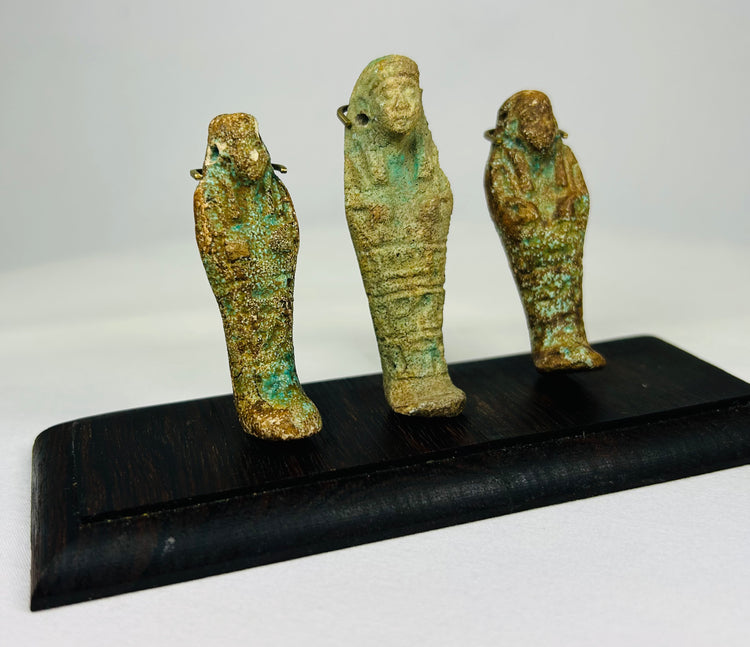 Egyptian ushabti trio with turquoise and ochre patina displayed on a dark wooden base against a white background.