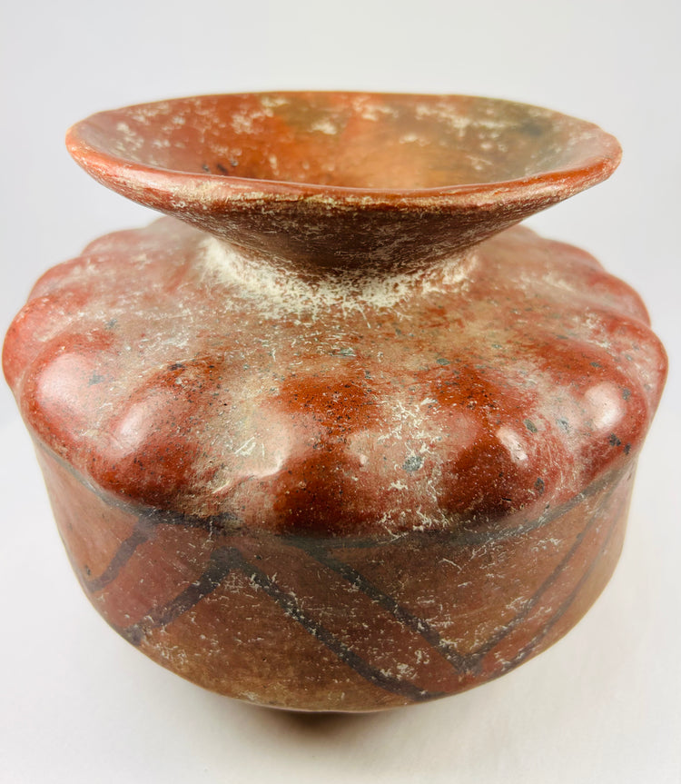 Pre-Columbian globular ceramic vessel with scalloped body and red-orange geometric painted designs on a white background.