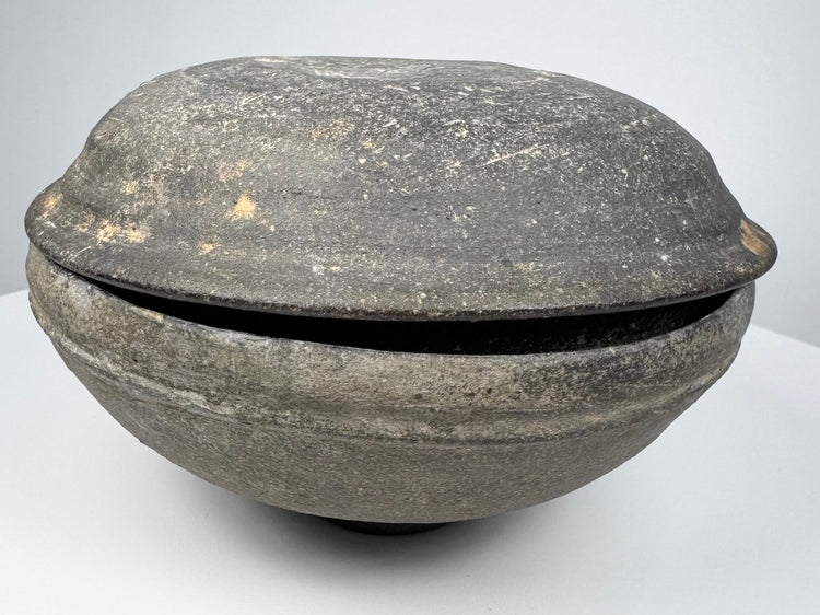 Side view of an ancient Chinese gray earthenware covered bowl with rounded lower body, fitted lid, and charcoal-gray patina.