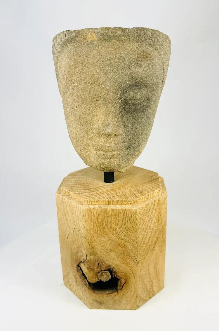 Khmer sandstone head fragment from the Angkor Period, 12th century Cambodia, depicting a serene deity or Buddha with downcast eyes and soft expression, mounted on a wooden display base, photographed on a white background.