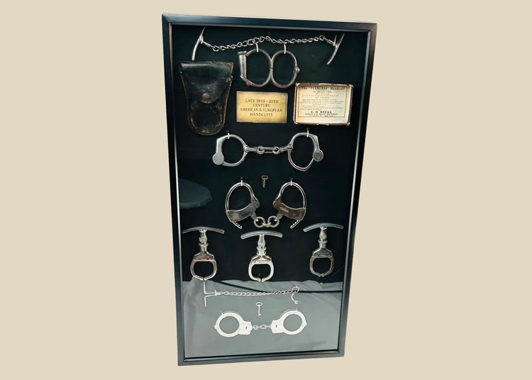 Framed shadow box display featuring a chronological collection of American and European handcuffs, leg irons, mechanical nippers, chain restraints, keys, and original leather pouches, mounted on black fabric with synthetic plaque, showcasing restraint devices from circa 1880s to 1950s.