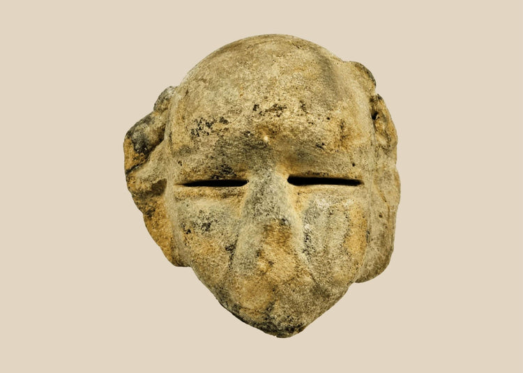 Ancient Mesoamerican stone mask fragment with minimalist facial features and narrow eye slits, carved from a single block of stone and mounted on a black wooden display stand, dating to 500 BCE–500 CE.