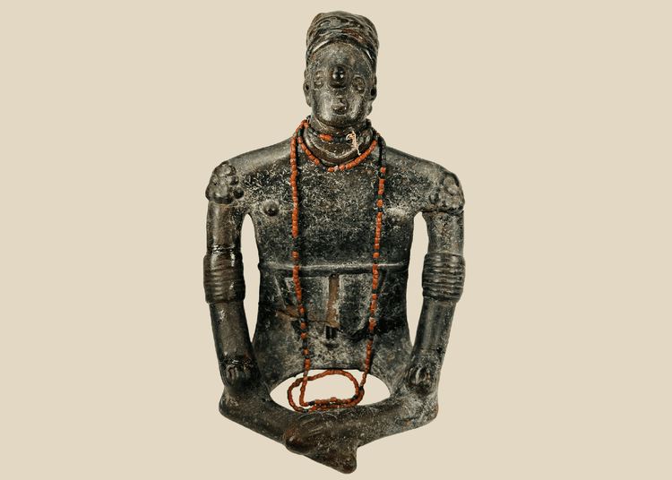 Front view of Pre-Columbian seated shamanic terracotta figure with red beads and headdress on a beige background.