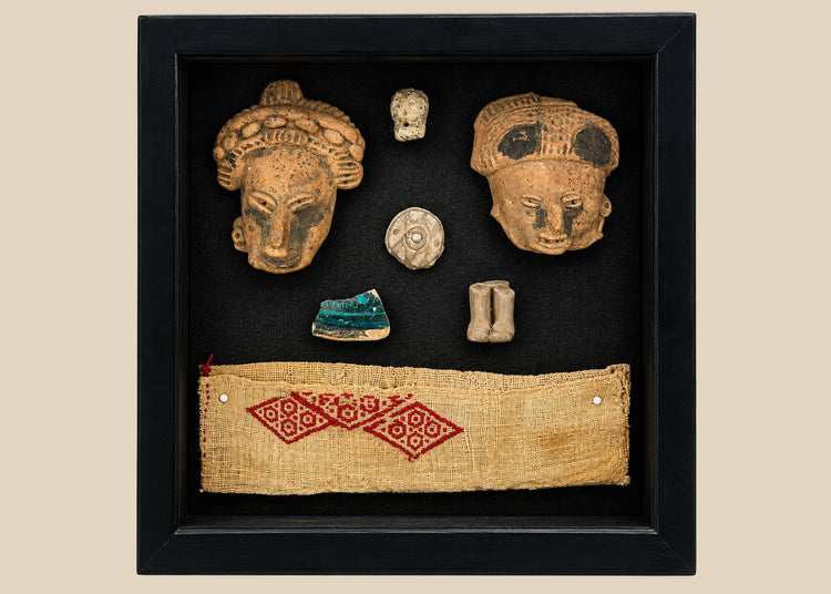 Framed display of Pre-Columbian artifacts including terracotta mask fragments, small figurines, carved ornaments, and a woven textile with red geometric pattern, set in a black shadowbox on a white background.