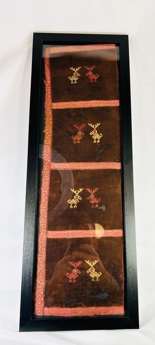 Ancient Pre-Columbian textile fragment featuring geometric animal motifs embroidered in red, gold, and tan thread on a dark brown woven ground, preserved in a protective display frame, dating to circa 1000–1400 CE, representing Andean artistry and symbolic ritual use.