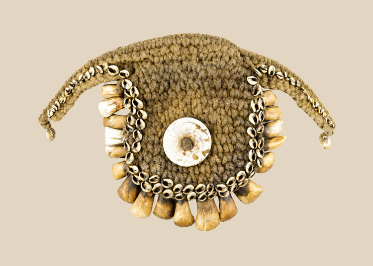 Antique ceremonial adornment from Papua New Guinea, late 19th–early 20th century, woven plant fiber with central cowrie shell and fringe of pierced animal teeth, displayed on a black stand.