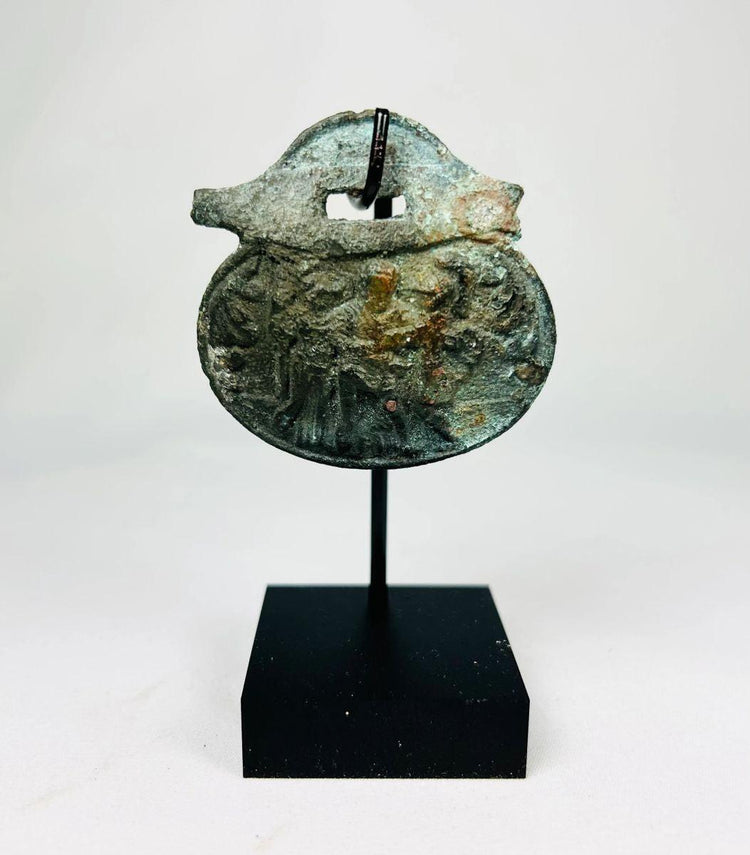 Ancient Roman bronze pendant, 1st–4th century CE, engraved with three standing figures including a helmeted figure, green patina, mounted on a custom stand.
