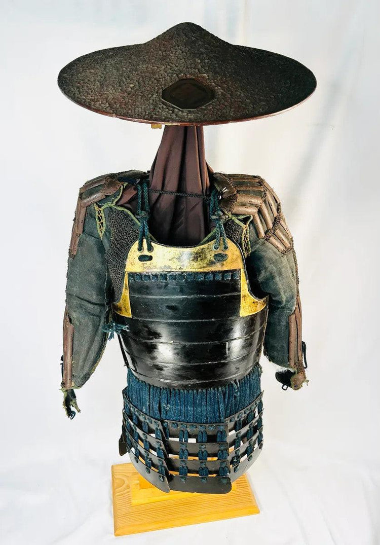 Authentic Japanese samurai armor from the Muromachi Period (1333–1573), featuring black-lacquered cuirass with gold accents, kote sleeves with chainmail reinforcement, dark-blue silk lacing, and a lacquered jingasa war hat bearing the Takeda clan crest, displayed on a wooden stand against a white background